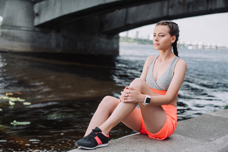 attractive sportswoman in sneakers sitting on quay near riverの写真素材