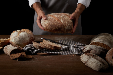 cropped image of male baker in apron holding bread over table with knife, bread and sackcloth