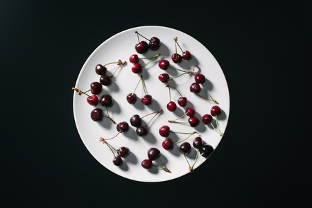 top view of fresh ripe red cherries on round white plate isolated on blackの写真素材