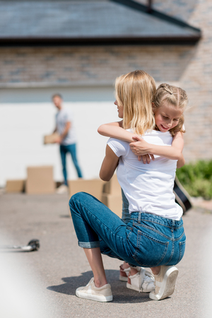 back view of woman hugging smiling daughter and man unpacking cardboard boxes behind in yard of new houseの写真素材