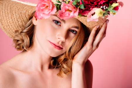 beautiful young woman in hat with flowers looking at camera isolated on pinkの写真素材