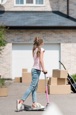 back view of kid riding on kick scooter near boxes and acoustic guitar in front of new houseの写真素材