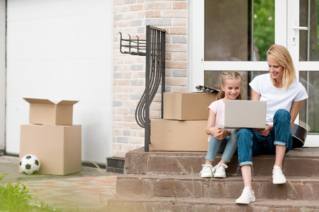 smiling woman with daughter using laptop on stairs with cardboard boxes and guitar in front of new...