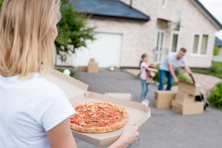partial view of  woman holding pizza while her husband and daughter unpacking cardboard boxes in front of new houseの写真素材