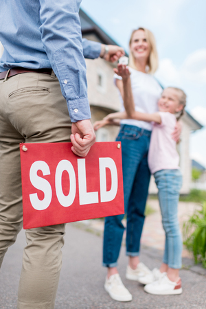 cropped image of realtor with sold sign giving key to young woman with daughter in front of new houseの写真素材