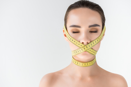 attractive young woman with closed mouth by measurement tape isolated on gray backgroundの写真素材