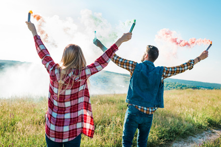 rear view of young couple holding colorful smoke bombs on rural meadowの写真素材