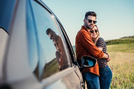 smiling stylish couple in sunglasses standing near car on rural meadowの写真素材