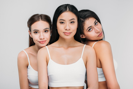 attractive multicultural girls in white bras posing together, isolated on greyの写真素材