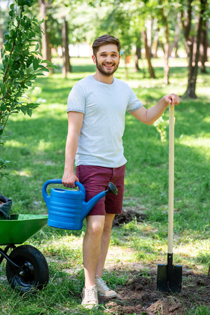 man with watering can and shovel standing in park with new trees in wheelbarrowの写真素材