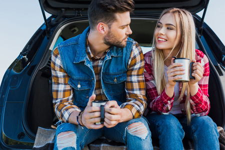 couple of young tourists with coffee cups sitting on car trunkの写真素材