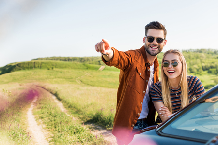 young stylish man pointing by hand to smiling woman near car on rural meadowの写真素材
