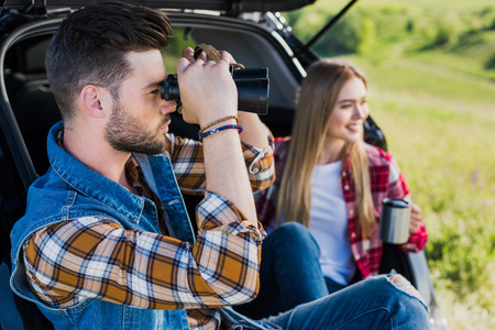 side view of male tourist looking through binoculars while his smiling girlfriend sitting near with...