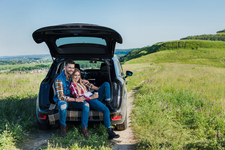 happy couple of stylish tourists with coffee cups sitting on car trunk in rural fieldの写真素材