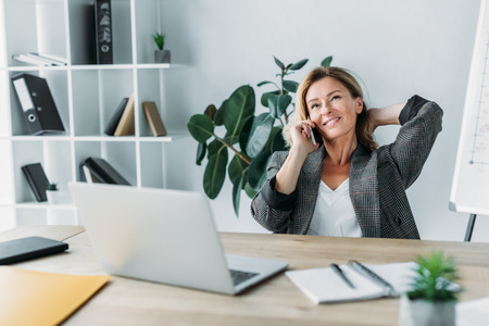 smiling attractive businesswoman talking by smartphone in office and sitting on chairの写真素材
