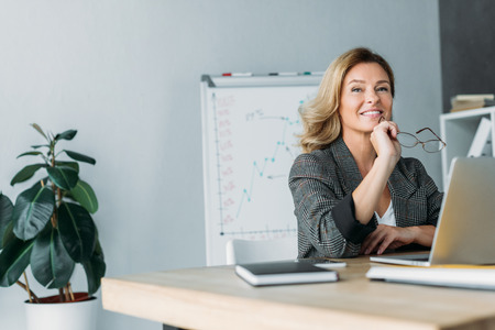 pensive smiling businesswoman holding glasses and looking away in officeの写真素材