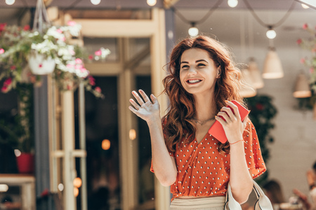 portrait of cheerful woman with smartphone in hand waiving to someone on streetの写真素材