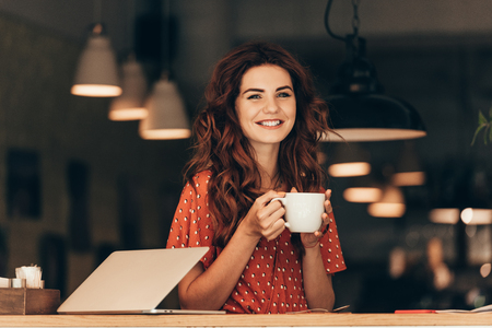 portrait of smiling woman with cup of coffee at table with laptop in cafeの写真素材