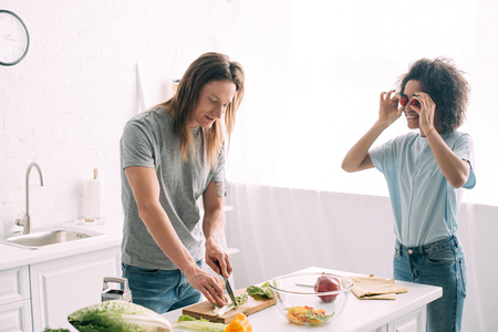 happy african american woman covering eyes by cherry tomatoes and boyfriend cutting salad leaves at kitchenの写真素材