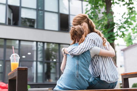 back view of mother and daughter hugging each other while resting on bench on streetの写真素材