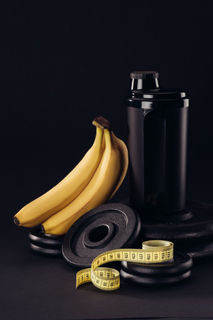 black protein shaker on pile of weight plates with bananas and measuring tape isolated on blackの写真素材