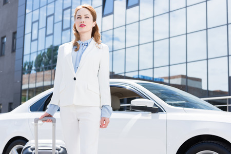 portrait of businesswoman in white suit with suitcase standing at car on streetの写真素材