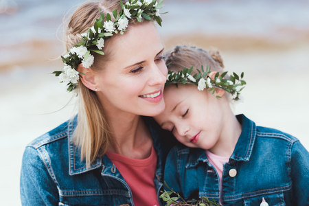 smiling mother and daughter in floral wreaths hugging on shoreの写真素材