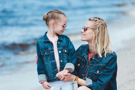 stylish mother and smiling daughter in sunglasses hugging on sea shoreの写真素材