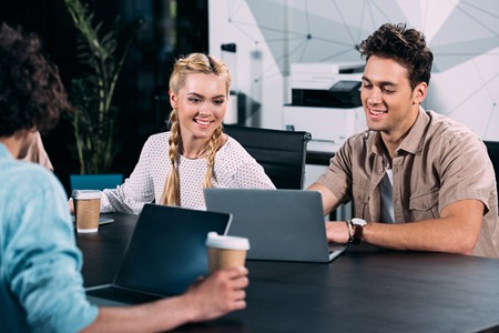 smiling businesspeople working at table with laptops and coffee cups at modern office