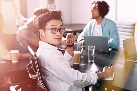 asian businessman in eyeglasses looking at camera while his partners having discussion at modern officeの写真素材