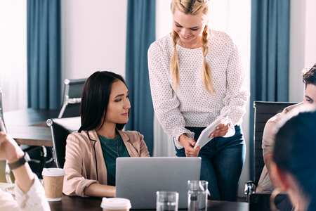 smiling businesswoman showing digital tablet to asian female colleague while other partners having discussion at modern officeの写真素材