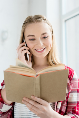 close-up portrait of happy teen student girl reading book and talking by phoneの写真素材