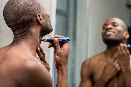 young bare-chested african american man shaving with electric trimmer in bathroomの写真素材