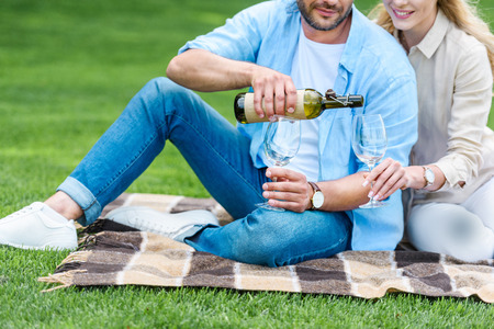 cropped shot of couple pouring wine while sitting on plaid at picnicの写真素材