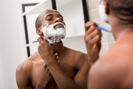 handsome african american man shaving with foam and razor and looking at mirrorの写真素材