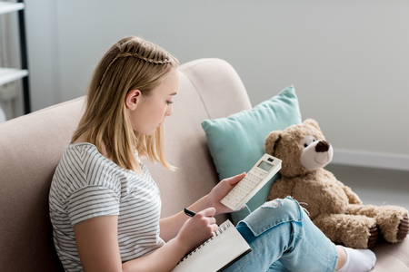 teen student girl writing in notebook and using calculator while sitting on couchの写真素材