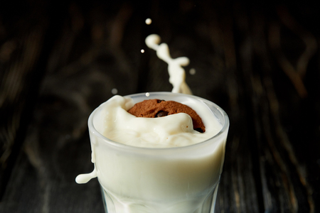 closeup view of chocolate cookie in milk glass with splashes on black wooden surfaceの写真素材