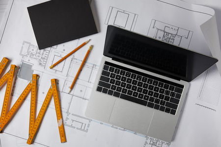 top view of laptop with blank screen, textbook, pencils and collapsible meter on blueprint