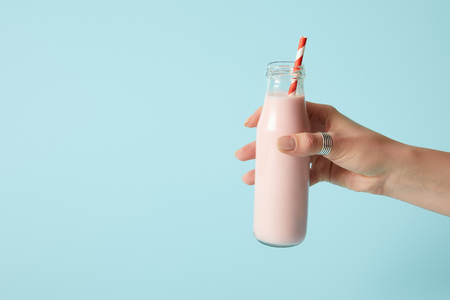 cropped shot of woman holding strawberry milkshake in bottle with drinking straw on blue backgroundの写真素材