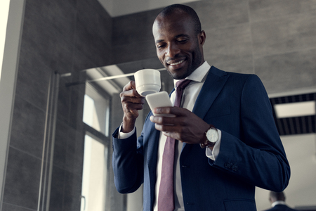 bottom view of smiling young businessman with cup of coffee using smartphone in bathroomの写真素材