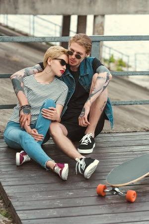 boyfriend with tattoos and stylish girlfriend sitting on bridge and hugging near skateboardの写真素材