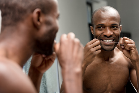 smiling young african american man holding dental floss and looking at mirror in bathroomの写真素材