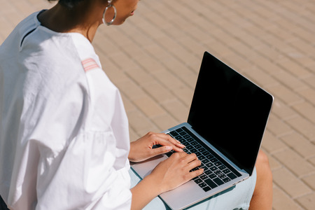 cropped image of african american businesswoman sitting on bench and using laptop on streetの写真素材