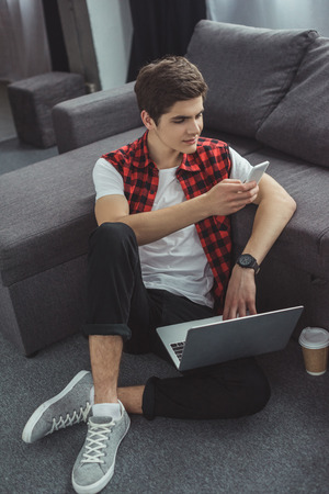 smiling teenager using smartphone and laptop while sitting on floor with coffeeの写真素材