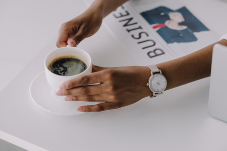 cropped image of african american businesswoman holding cup of coffee at table with newspaper in officeの写真素材