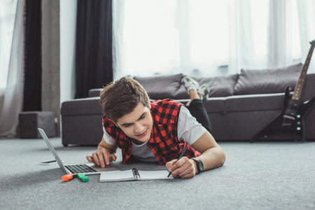 teen boy studying with copybook and laptop while lying on floorの写真素材