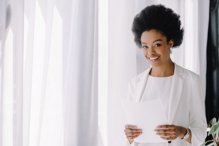 attractive smiling african american businesswoman holding documents in officeの写真素材