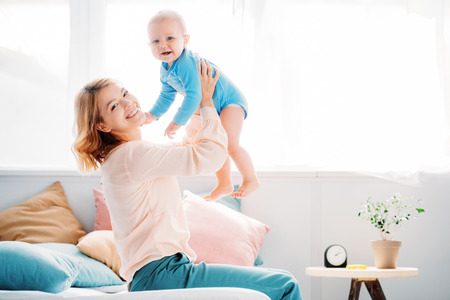 side view of smiling mother raising laughing little child while sitting on bed at home
