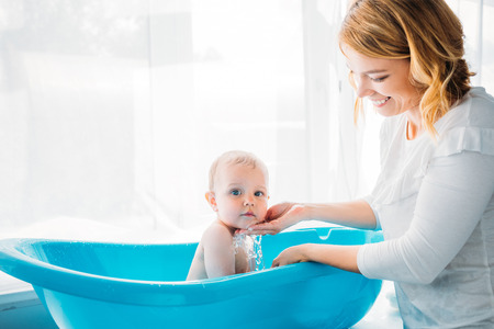 side view of happy mother washing her adorable little child in plastic baby bathtub at home
