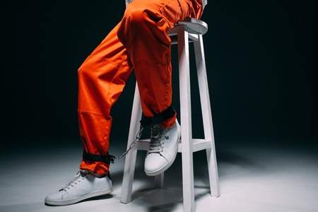 Cropped view of man in orange uniform with cuffs on legs sitting on stool on dark backgroundの写真素材